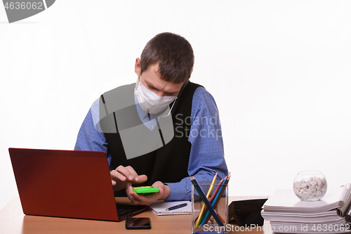 Image of An office employee in a protective medical mask is talking on the phone, counting on a calculator