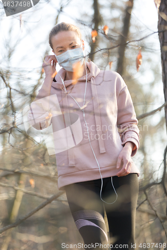 Image of Portrait of caucasian sporty woman wearing medical protection face mask while walking in park, relaxing and listening to music. Corona virus, or Covid-19, is spreading all over the world