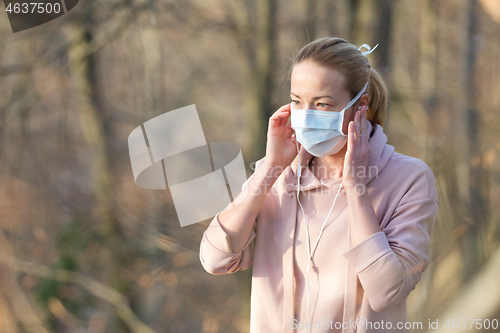 Image of Portrait of caucasian sporty woman wearing medical protection face mask while walking in park, relaxing and listening to music. Corona virus, or Covid-19, is spreading all over the world