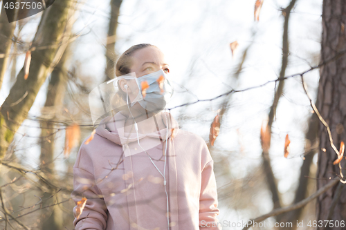 Image of Portrait of caucasian sporty woman wearing medical protection face mask while walking in park, relaxing and listening to music. Corona virus, or Covid-19, is spreading all over the world