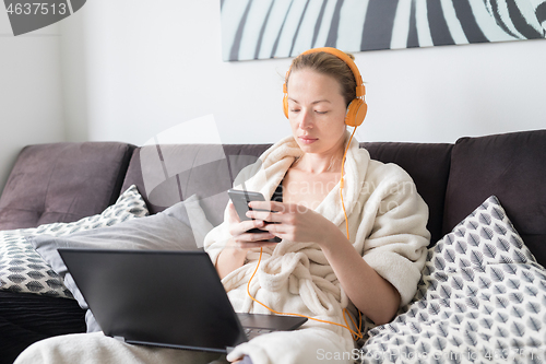 Image of Social distancing. Stay at home. Woman in bathrobe being comfortable at her home sofa, using social media apps on phone for video chatting and stying connected with her loved ones.