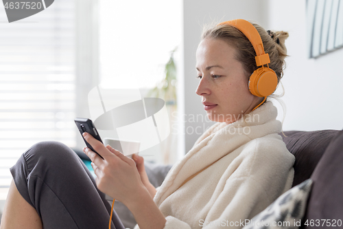 Image of Stay at home. Social distancing. Woman at home relaxing on sofa couch drinking tea from white cup, listening to relaxing music, stay connected to friens and family via social networks on mobile.