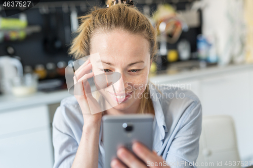 Image of Young smiling cheerful pleased woman indoors at home kitchen using social media apps on mobile phone for chatting and stying connected with her loved ones. Stay at home, social distancing lifestyle.