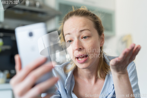 Image of Young smiling cheerful pleased woman indoors at home kitchen using social media apps on mobile phone for chatting and stying connected with her loved ones. Stay at home, social distancing lifestyle.