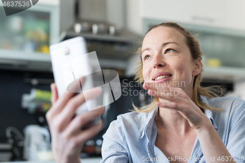 Image of Young smiling cheerful pleased woman indoors at home kitchen using social media apps on mobile phone for chatting and stying connected with her loved ones. Stay at home, social distancing lifestyle.