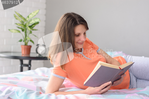 Image of Girl at home lying in bed reads a book.