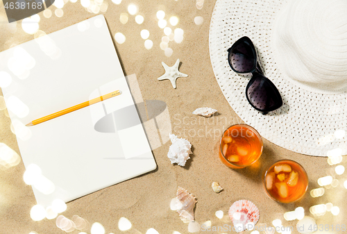 Image of notebook, cocktails, hat and shades on beach sand