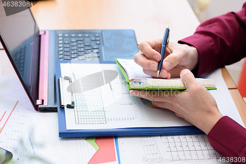 Image of a person writes in a notebook data from a laptop and documents laid out on the table