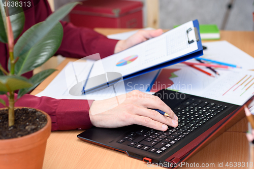 Image of close-up of hands typing on a laptop on a desk with documents, graphs and tables
