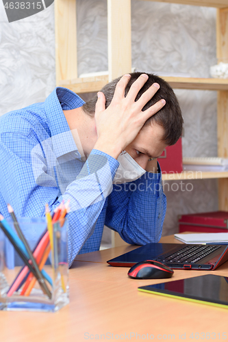 Image of A man sits at a desk holding his head in a medical mask