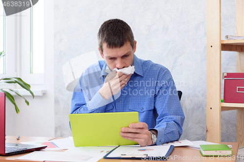 Image of A man in the office with a tablet in his hand coughs in a napkin