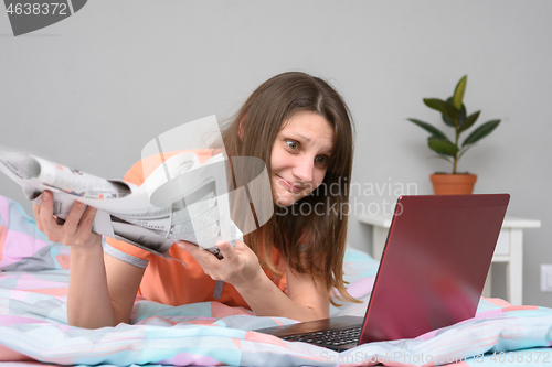 Image of A girl with newspapers in her hands lies in bed and looks at the laptop screen in search of work