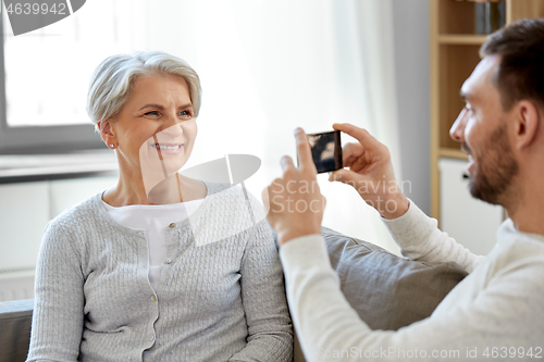 Image of adult son photographing senior mother at home