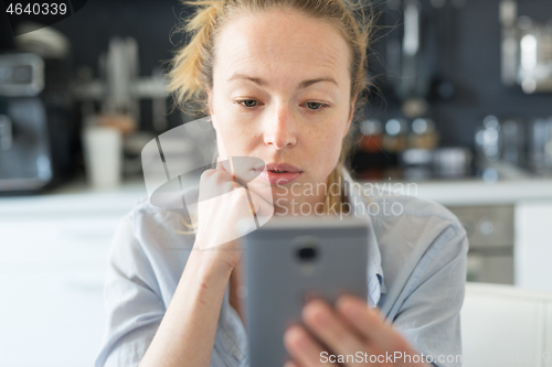 Image of Young smiling cheerful pleased woman indoors at home kitchen using social media apps on mobile phone for chatting and stying connected with her loved ones. Stay at home, social distancing lifestyle.