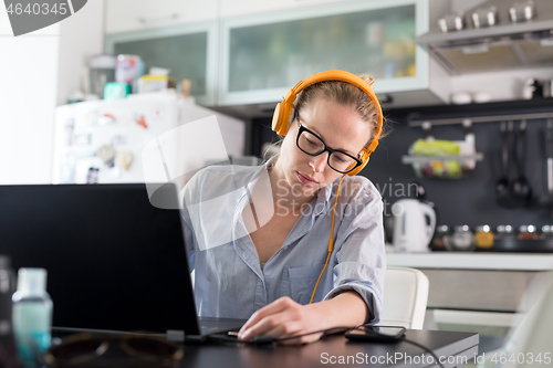 Image of Female freelancer in her casual home clothing working remotly from her dining table in the morning. Home kitchen in the background.