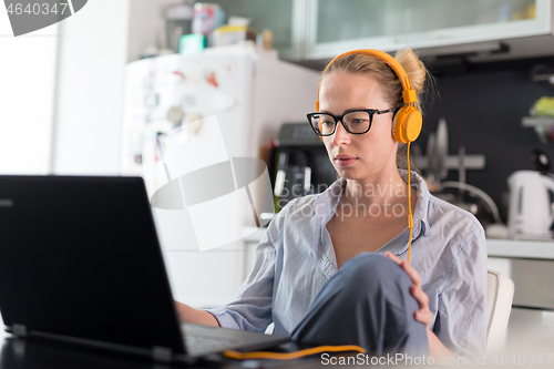 Image of Female freelancer in her casual home clothing working remotly from her dining table in the morning. Home kitchen in the background.