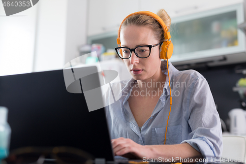 Image of Female freelancer in her casual home clothing working remotly from her dining table in the morning. Home kitchen in the background.