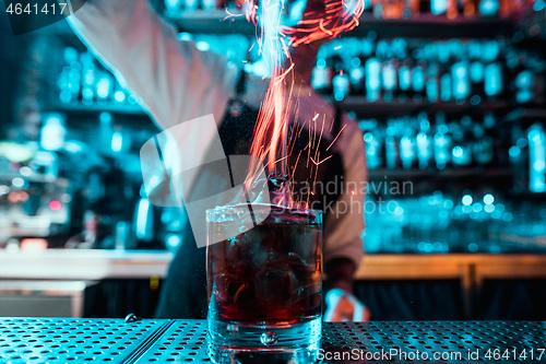 Image of Glass of fiery cocktail on the bar counter against the background of bartenders hands with fire