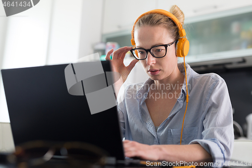 Image of Female freelancer in her casual home clothing working remotly from her dining table in the morning. Home kitchen in the background.