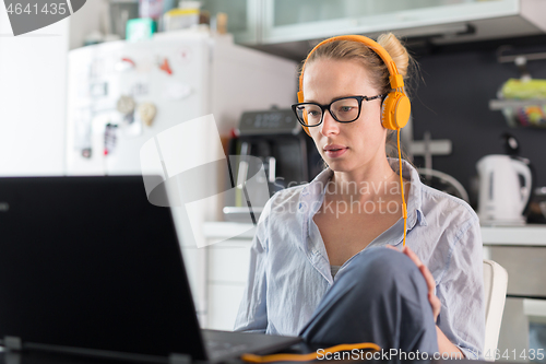 Image of Female freelancer in her casual home clothing working remotly from her dining table in the morning. Home kitchen in the background.