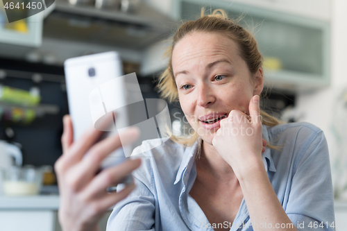 Image of Young smiling cheerful pleased woman indoors at home kitchen using social media apps on mobile phone for chatting and stying connected with her loved ones. Stay at home, social distancing lifestyle.