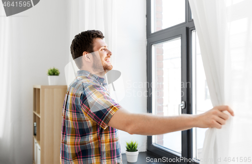 Image of young man opening window curtain at home