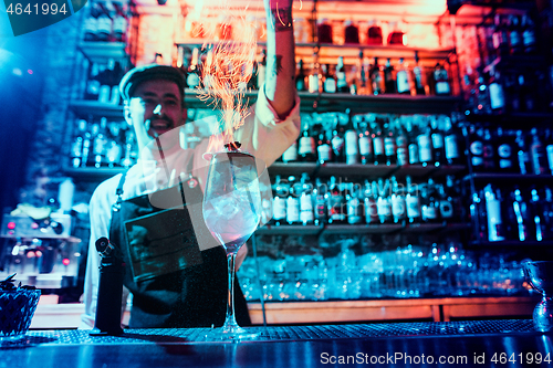 Image of Glass of fiery cocktail on the bar counter against the background of bartenders hands with fire