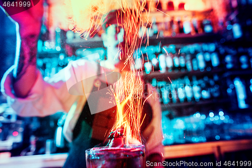 Image of Glass of fiery cocktail on the bar counter against the background of bartenders hands with fire