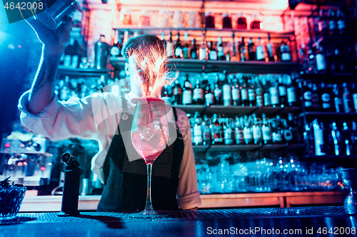 Image of Glass of fiery cocktail on the bar counter against the background of bartenders hands with fire