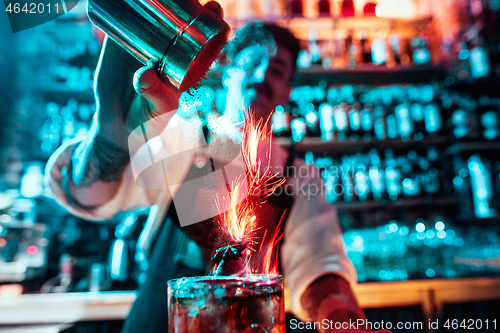 Image of Glass of fiery cocktail on the bar counter against the background of bartenders hands with fire