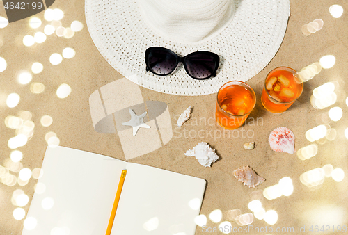 Image of notebook, cocktails, hat and shades on beach sand