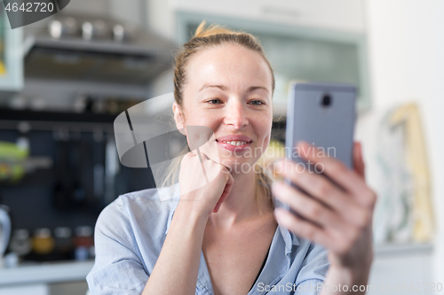 Image of Young smiling cheerful pleased woman indoors at home kitchen using social media apps on mobile phone for chatting and stying connected with her loved ones. Stay at home, social distancing lifestyle.