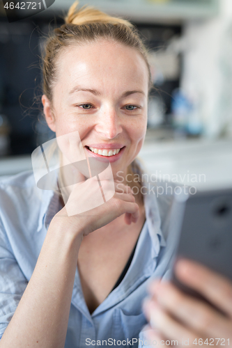 Image of Young smiling cheerful pleased woman indoors at home kitchen using social media apps on mobile phone for chatting and stying connected with her loved ones. Stay at home, social distancing lifestyle.