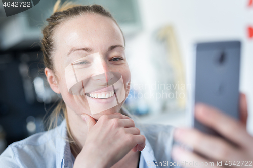 Image of Young smiling cheerful pleased woman indoors at home kitchen using social media apps on mobile phone for chatting and stying connected with her loved ones. Stay at home, social distancing lifestyle.