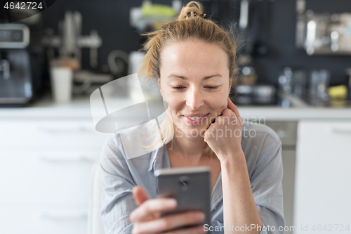 Image of Young smiling cheerful pleased woman indoors at home kitchen using social media apps on mobile phone for chatting and stying connected with her loved ones. Stay at home, social distancing lifestyle.