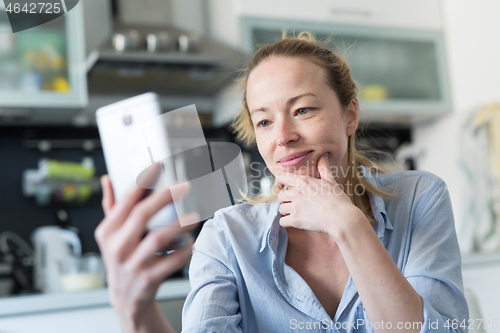 Image of Young smiling cheerful pleased woman indoors at home kitchen using social media apps on mobile phone for chatting and stying connected with her loved ones. Stay at home, social distancing lifestyle.