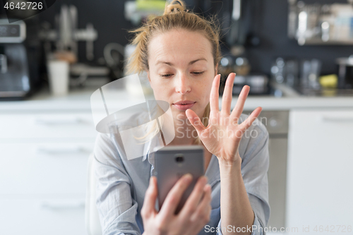 Image of Young pleased woman indoors at home kitchen using social media apps on mobile phone for chatting and stying connected with her loved ones. Stay at home, social distancing lifestyle.