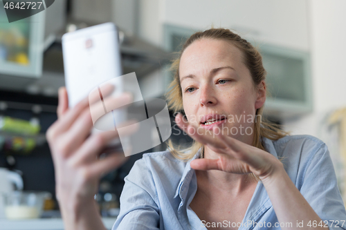 Image of Young pleased woman indoors at home kitchen using social media apps on mobile phone for chatting and stying connected with her loved ones. Stay at home, social distancing lifestyle.