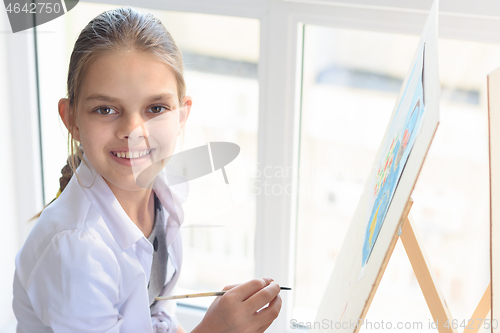Image of Happy teenager girl draws on an easel by the window and looked into the frame with a smile
