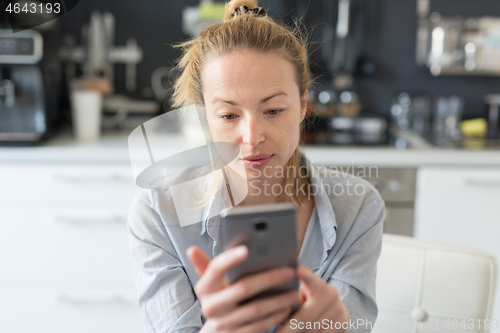 Image of Worried woman indoors at home kitchen using social media apps on phone for video chatting and stying connected with her loved ones. Stay at home, social distancing lifestyle