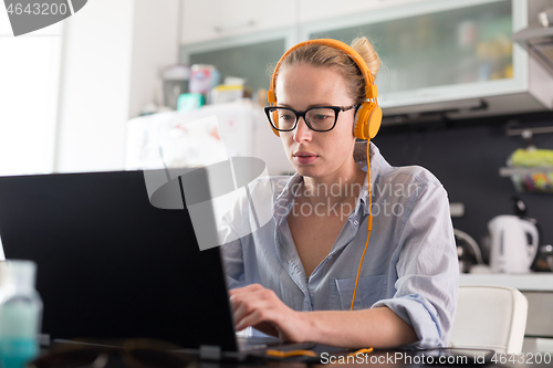 Image of Female freelancer in her casual home clothing working remotly from her dining table in the morning. Home kitchen in the background.