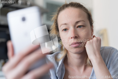 Image of Worried woman indoors at home kitchen using social media apps on phone for video chatting and stying connected with her loved ones. Stay at home, social distancing lifestyle