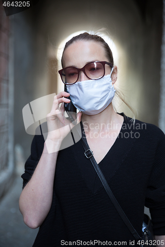 Image of COVID-19 pandemic coronavirus. Casual caucasian woman at medieval city street using mobile phone, wearing protective face mask against spreading of coronavirus and disease transmission