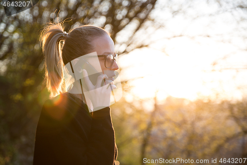 Image of Backlit rear view of young woman talking on cell phone outdoors in park at sunset. Girl holding mobile phone, using digital device, looking at setting sun