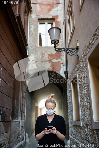 Image of COVID-19 pandemic coronavirus. Casual caucasian woman at medieval city street using mobile phone, wearing protective face mask against spreading of coronavirus and disease transmission