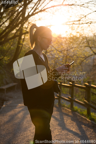 Image of Backlit rear view of young woman talking on cell phone outdoors in park at sunset. Girl holding mobile phone, using digital device, looking at setting sun