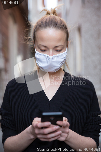 Image of COVID-19 pandemic coronavirus. Casual caucasian woman at medieval city street using mobile phone, wearing protective face mask against spreading of coronavirus and disease transmission