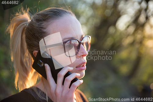 Image of Backlit rear view of young woman talking on cell phone outdoors in park at sunset. Girl holding mobile phone, using digital device, looking at setting sun