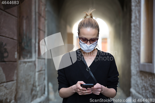 Image of COVID-19 pandemic coronavirus. Casual caucasian woman at medieval city street using mobile phone, wearing protective face mask against spreading of coronavirus and disease transmission