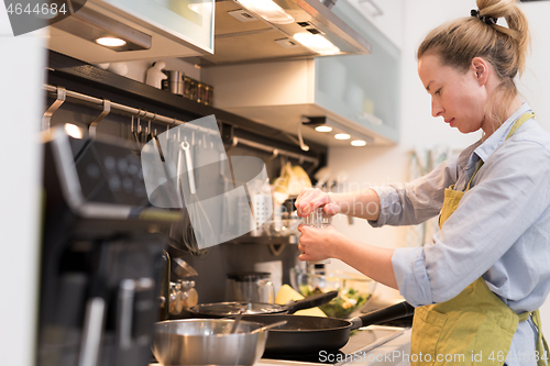 Image of Stay at home housewife woman cooking in kitchen, salting dish in a saucepan, preparing food for family dinner.